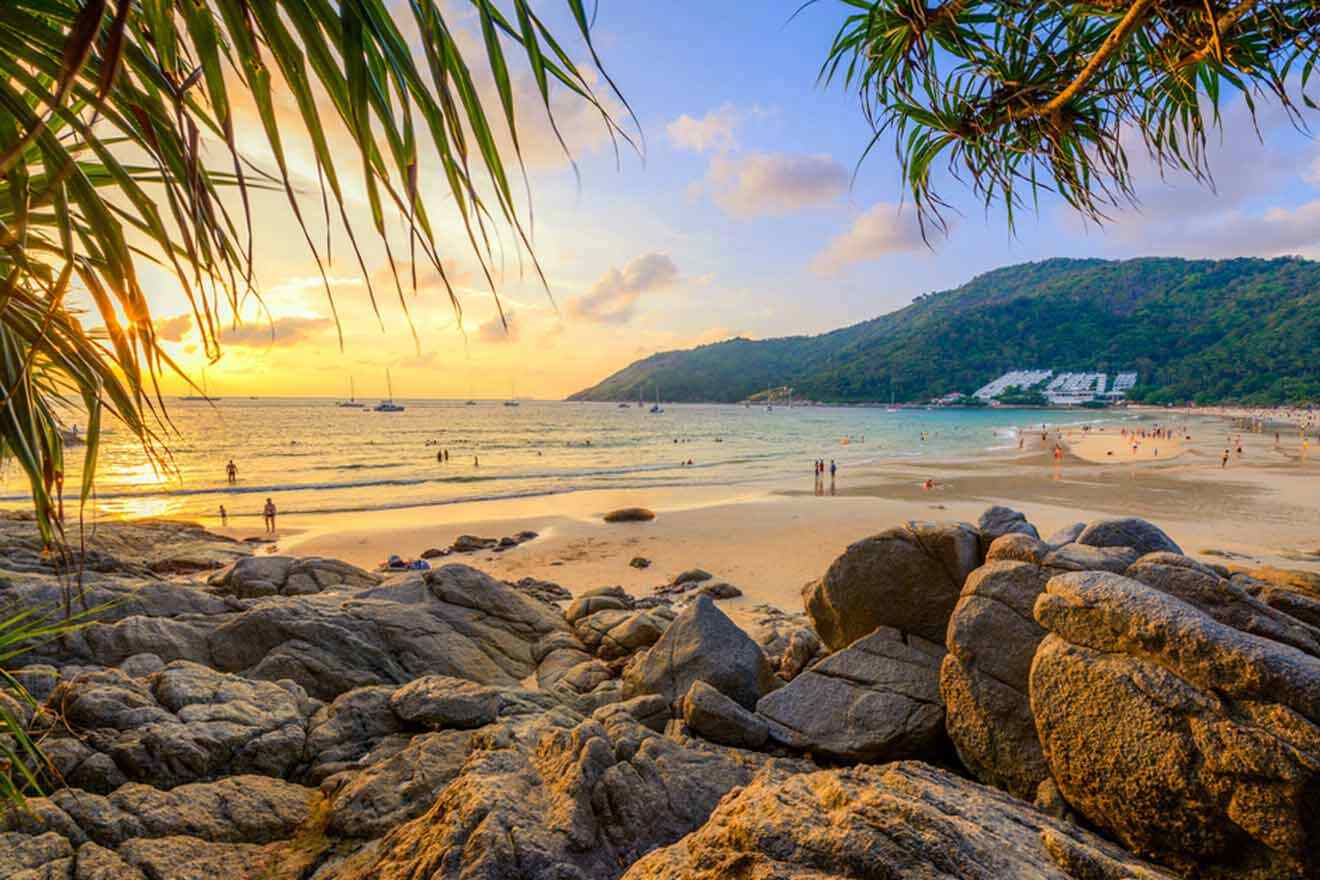 Golden sunset over Nai Harn Beach with visitors strolling along the shore and sailboats in the distance, framed by tropical palm leaves and rocks, embodying the peaceful beauty of Phuket's coastline.