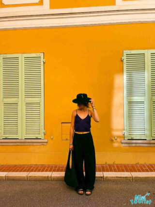 The writer of the post in a stylish outfit stands against a vibrant yellow building with green shutters in Menton, France,