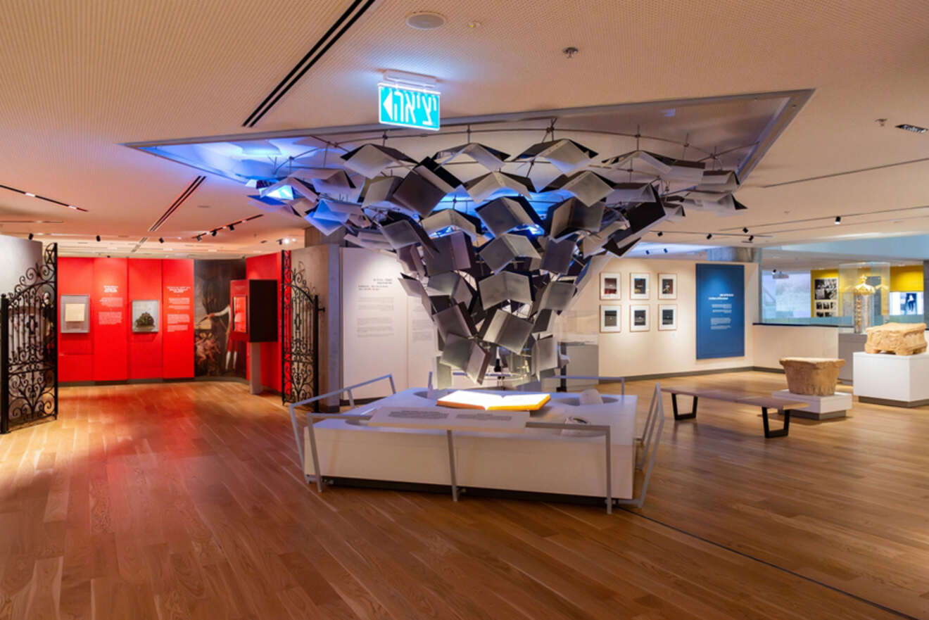 A modern museum gallery with an intricate ceiling installation composed of geometric shapes, surrounded by various exhibits and informational displays