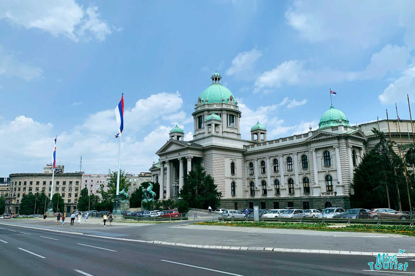 The National Assembly of Serbia in Belgrade, showcasing neoclassical architecture against a blue sky, flanked by Serbian flags.