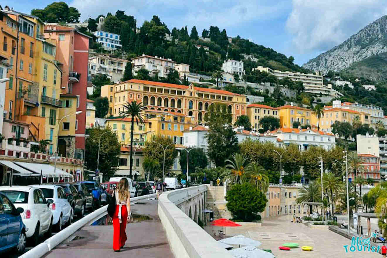 The writer of the post walks across the bridge in the seaside town of Menton, France, with a backdrop of colorful buildings and the Mediterranean coastline