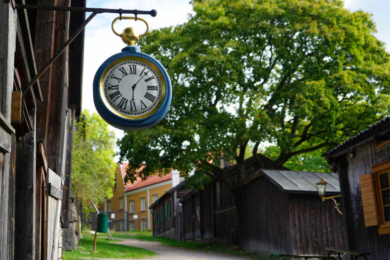 Vintage blue street clock hanging on a dark wooden beam with a historical alleyway in the background in Luostarinmäki, Turku