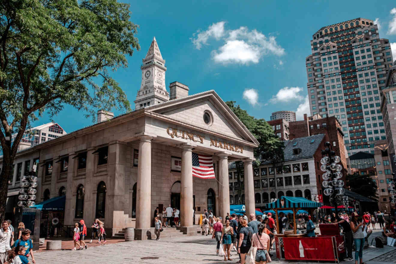 Quincy Market in Boston bustling with visitors and flanked by green trees, with the Custom House Tower visible in the background under a clear blue sky