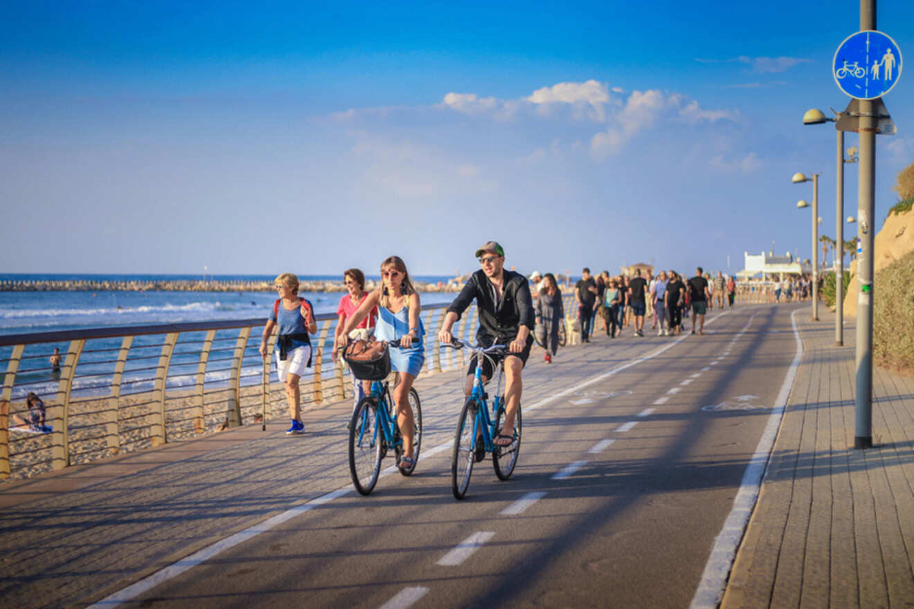 People cycling and walking along a bustling beachfront promenade with a clear blue sky overhead and calm sea in the background