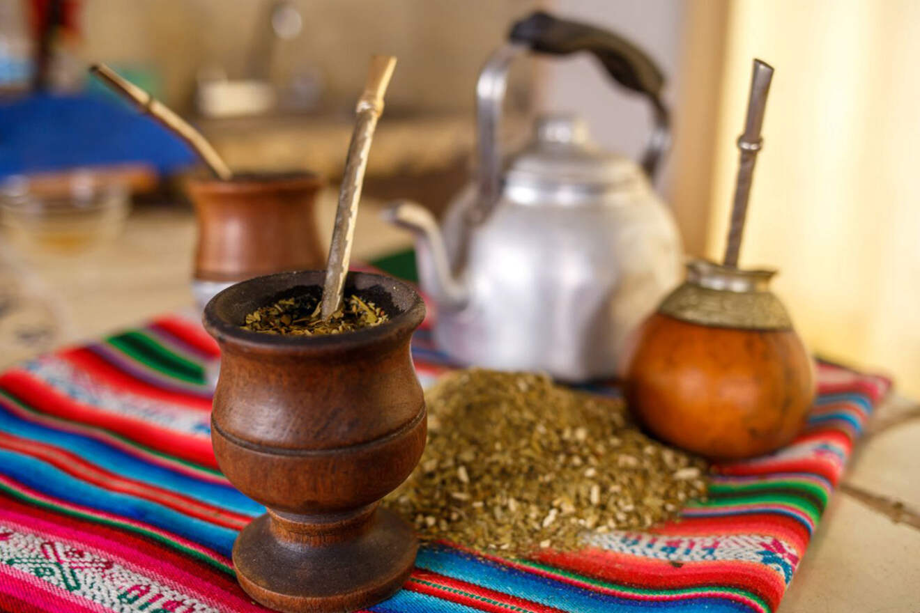 A close-up of a traditional Argentine yerba mate setup with a wooden mate cup, bombilla (metal straw), and a kettle, set against a colorful woven fabric