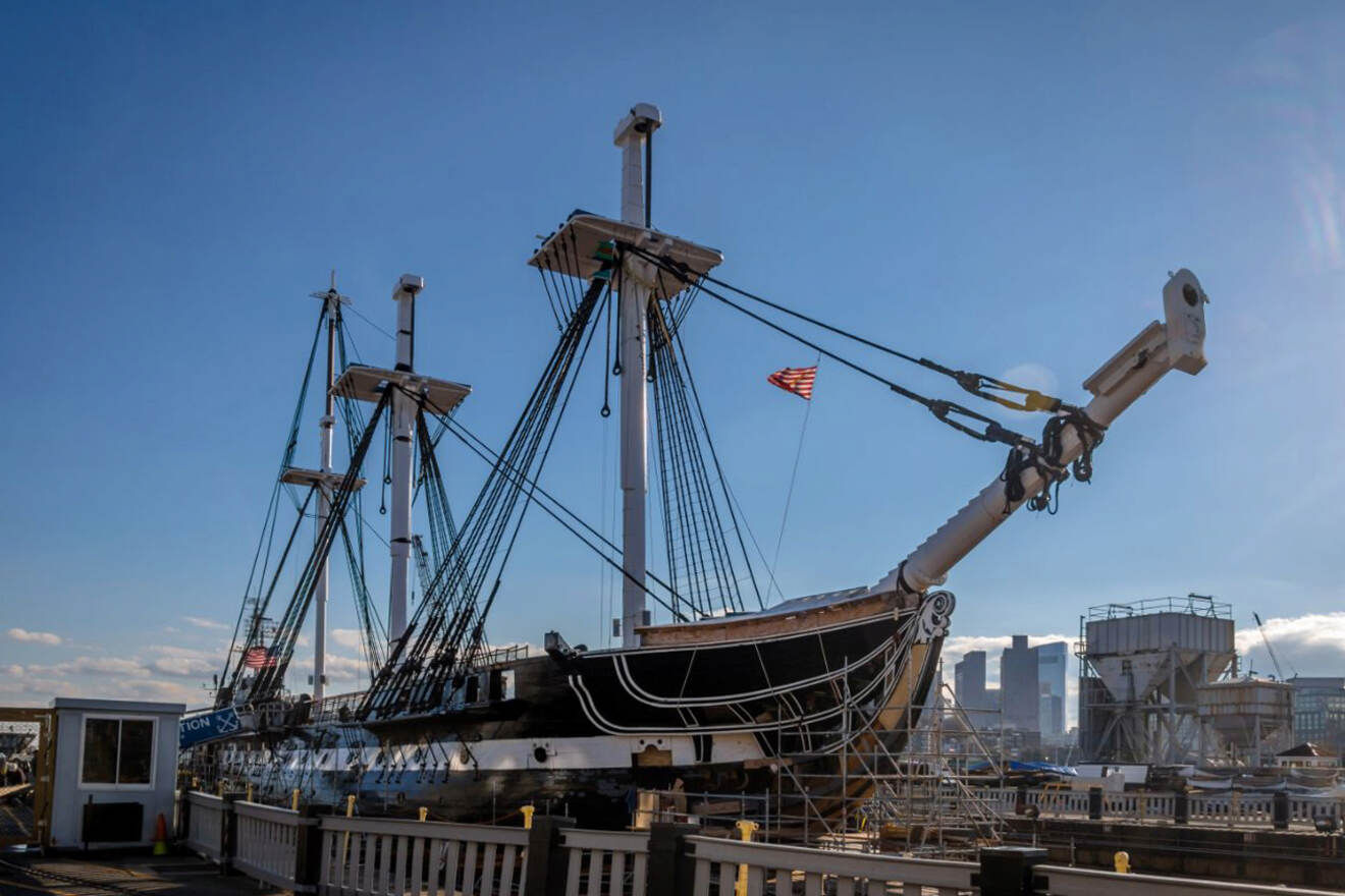 The USS Constitution, an iconic historic warship docked at the Charlestown Navy Yard in Boston, with modern buildings in the distance under a blue sky