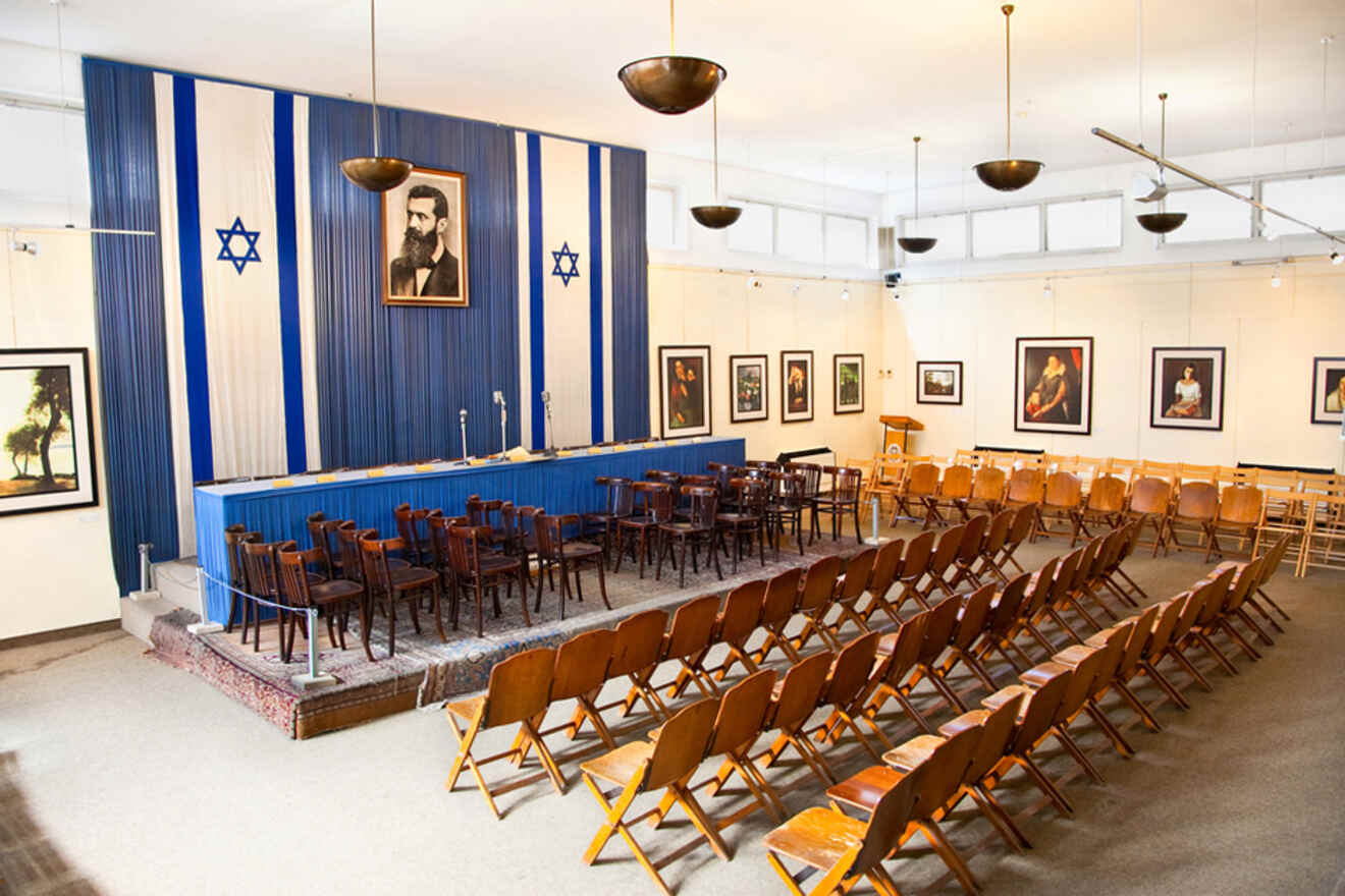 An interior view of a ceremonial hall with rows of wooden chairs facing a stage adorned with the Israeli flag and portraits