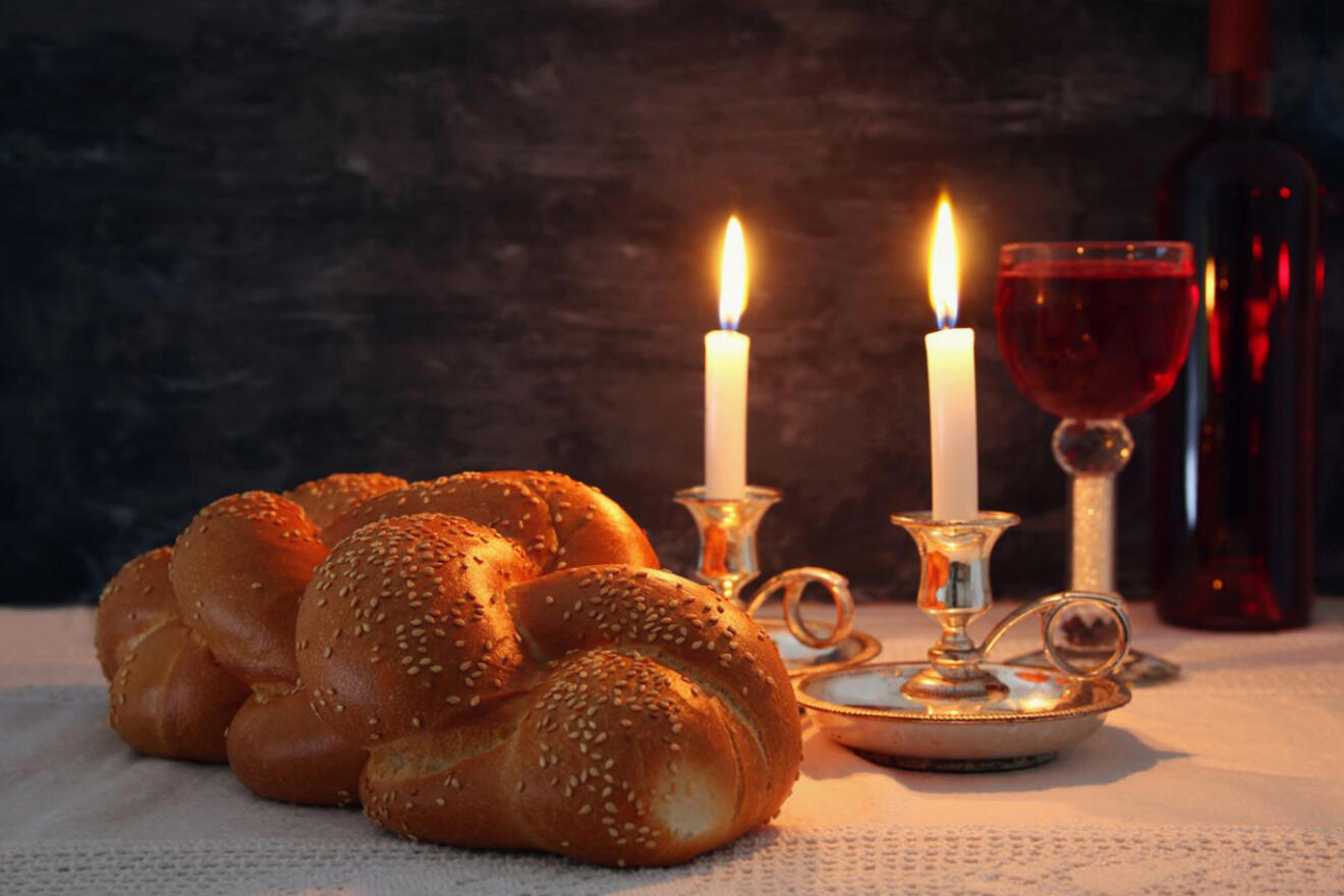 Traditional Jewish Shabbat table setting featuring two lit candles, challah bread, wine in a glass and bottle, and silver candlesticks