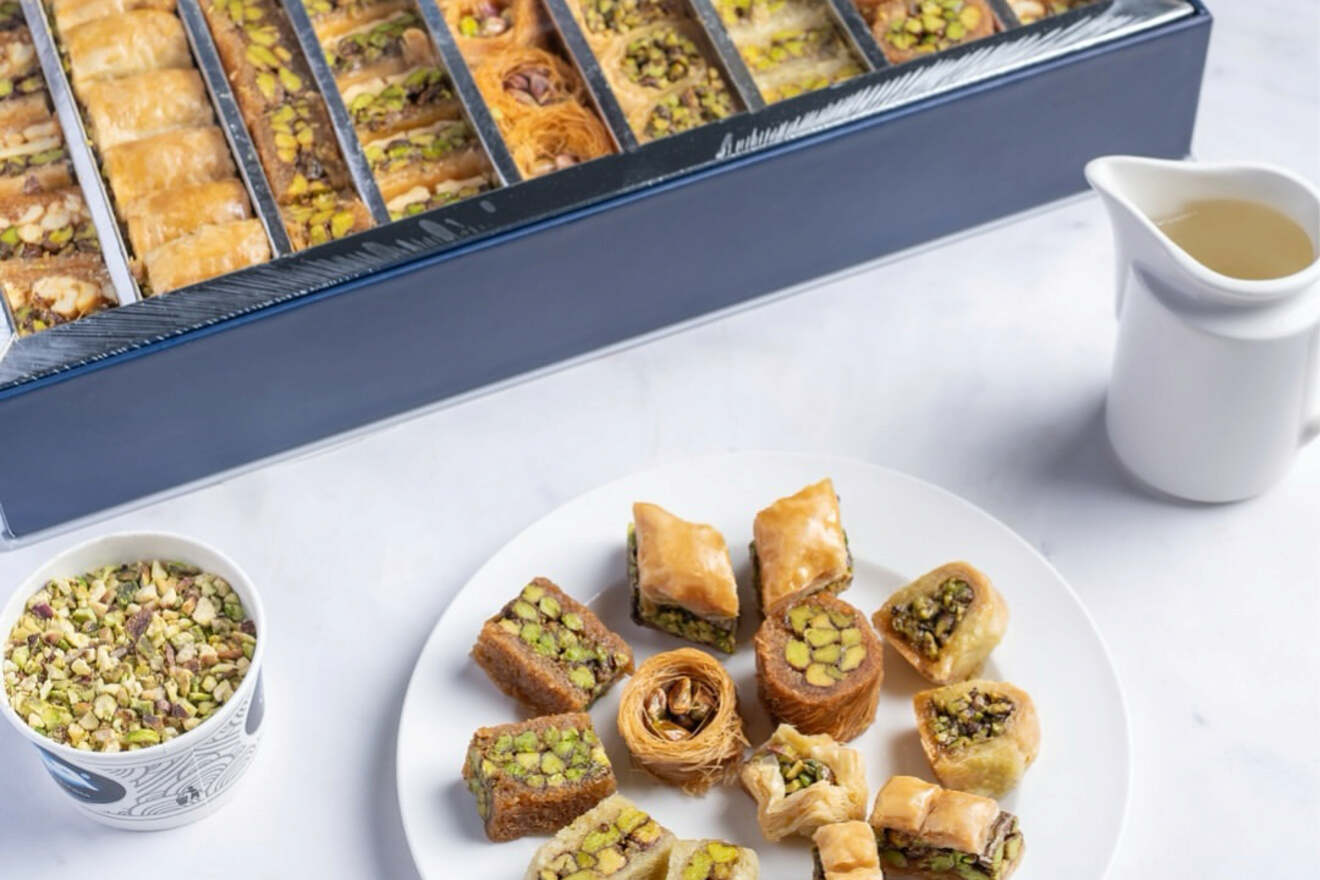A selection of Middle Eastern desserts, including baklava, arranged elegantly on a white plate with a box full of assorted treats and a jug of syrup on the side