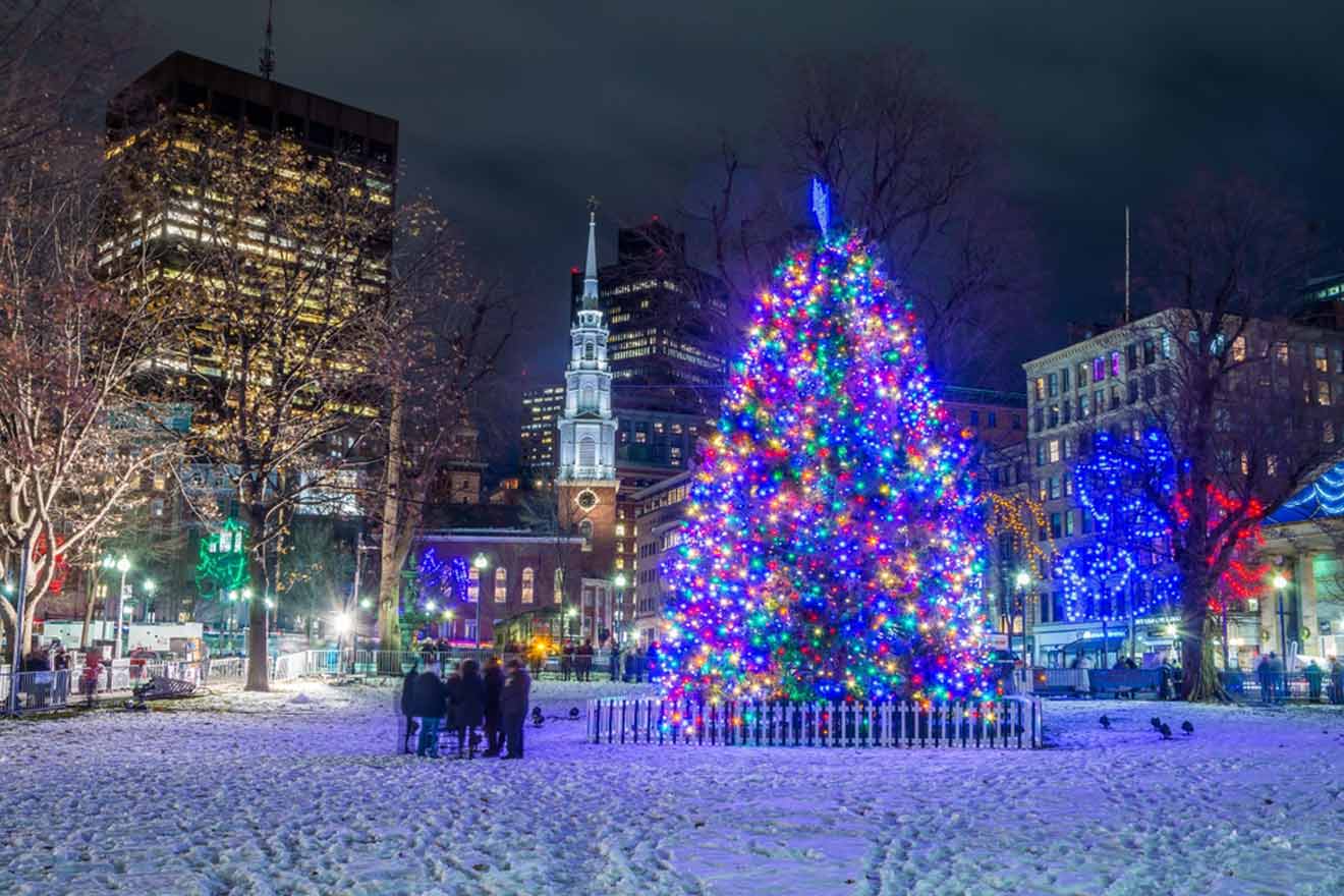 A vibrant Christmas tree illuminated with multicolored lights stands out in a snowy urban park at night, with silhouettes of people gathered nearby and city buildings in the backdrop.