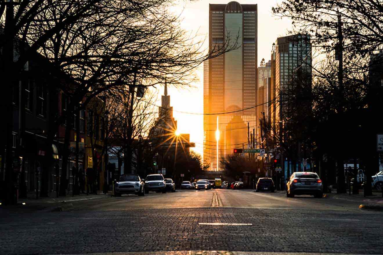 Sunset view of a Dallas street in Deep Ellum with the sun casting a warm glow between high-rise buildings, creating a picturesque urban scene.