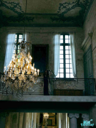A majestic chandelier illuminates a woman on a balcony inside the opulent Musée Calvet, with intricate frescoes and grand windows