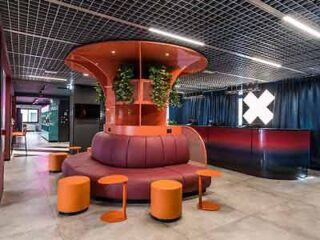 A modern lounge area with vibrant orange seating, a unique red mushroom-shaped centerpiece, surrounded by plants and under a geometric ceiling design.
