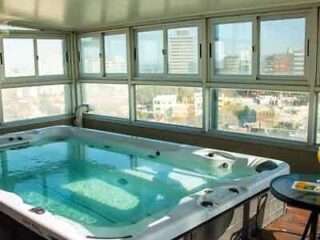 An enclosed rooftop spa area with a large jacuzzi, surrounded by windows offering a panoramic view of the urban landscape.