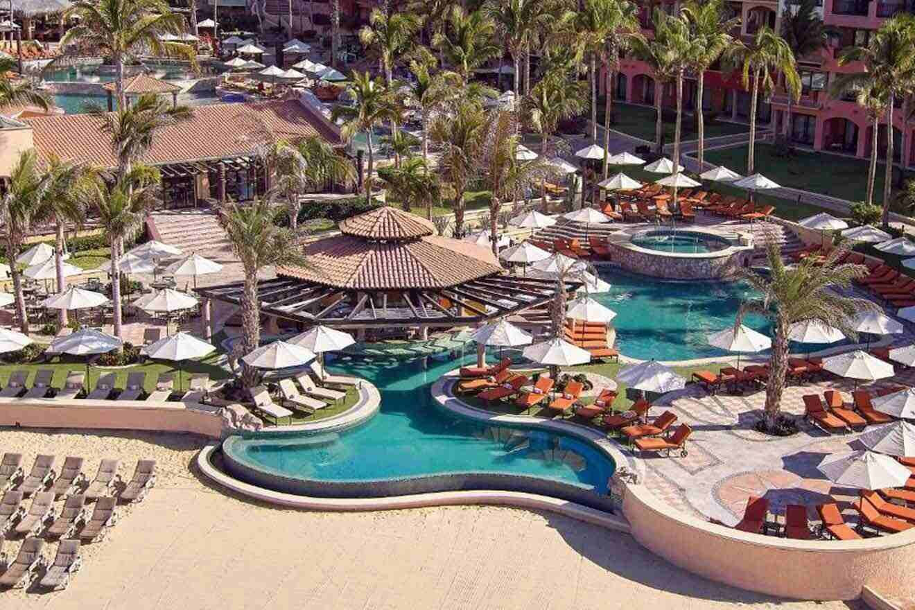 Aerial view of a family-friendly resort in Cabo San Lucas with multiple swimming pools, sun loungers, umbrellas, and palm trees, surrounded by pink-hued buildings.