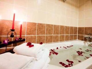 A romantic hotel bathtub setup with rose petals scattered in the water and over the adjacent towels, accompanied by a red candle, setting the mood for a relaxing and intimate evening.