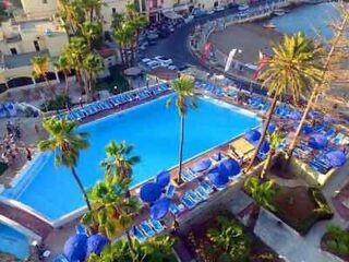 An aerial view of a hotel pool area with blue sun loungers and palm trees, adjacent to a beachside promenade.