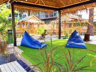A serene garden area with blue bean bags on the lawn under a rustic thatched-roof pavilion, providing a casual and relaxing outdoor lounge space amidst tropical plants.