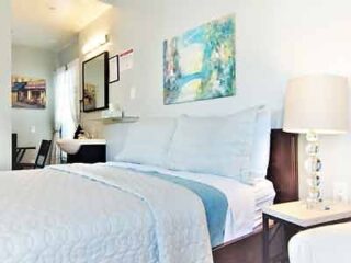 A bright hotel room with a queen bed, white bedding, modern lighting, and colorful artwork on the walls.