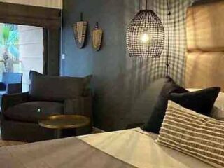 A modern hotel room with a comfortable bed, gray armchair, wooden bedside table, and stylish hanging light fixtures.
