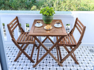 Uma pequena mesa de madeira com duas cadeiras em uma varanda azulejada, com duas xícaras de café, um prato de doces e um vaso de manjericão.