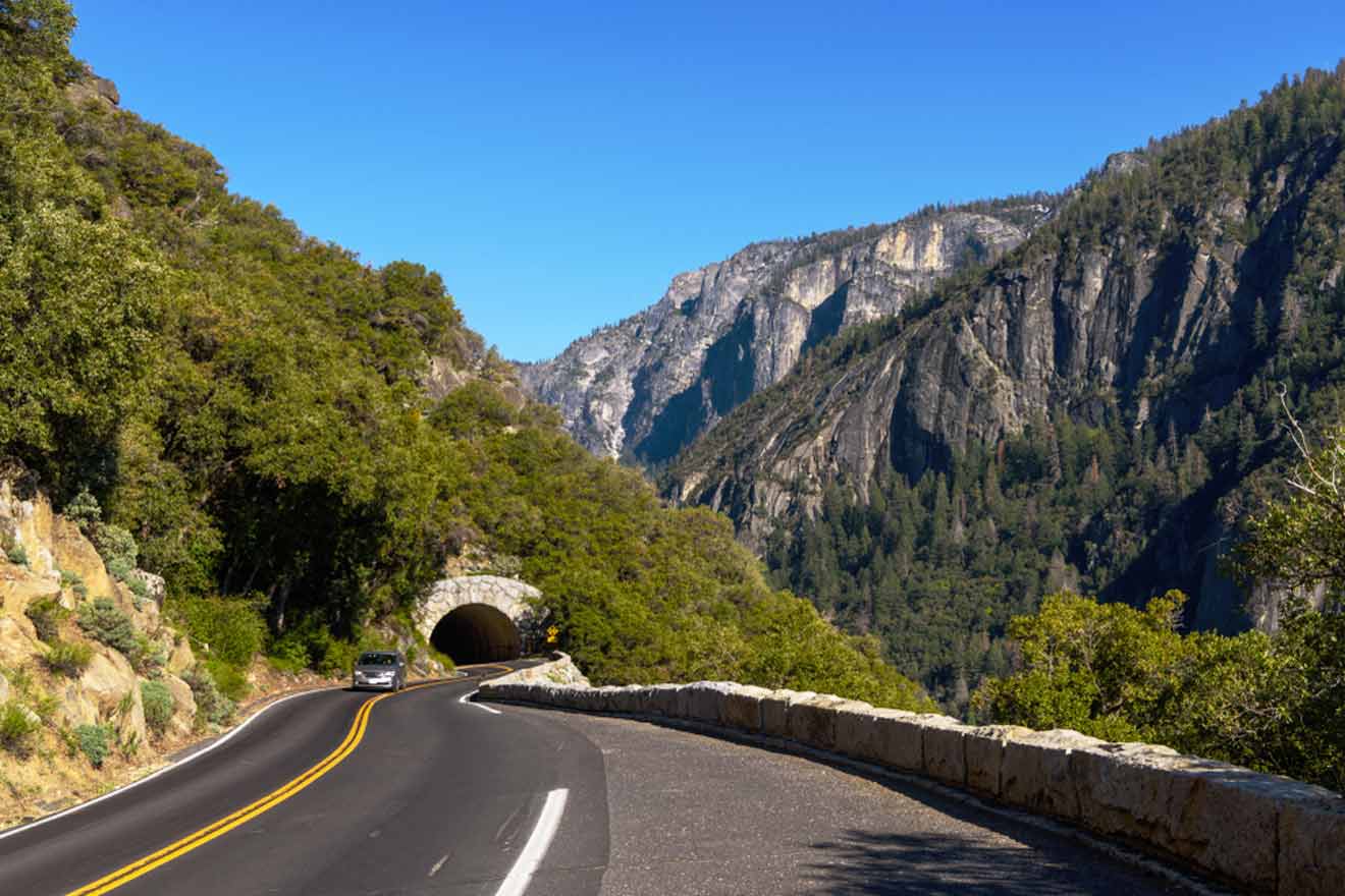 The winding road to Big Oak Flat Entrance in Yosemite National Park with a clear blue sky overhead. A stone tunnel carved through a hill awaits travelers, surrounded by steep, forested slopes and rugged mountain scenery.