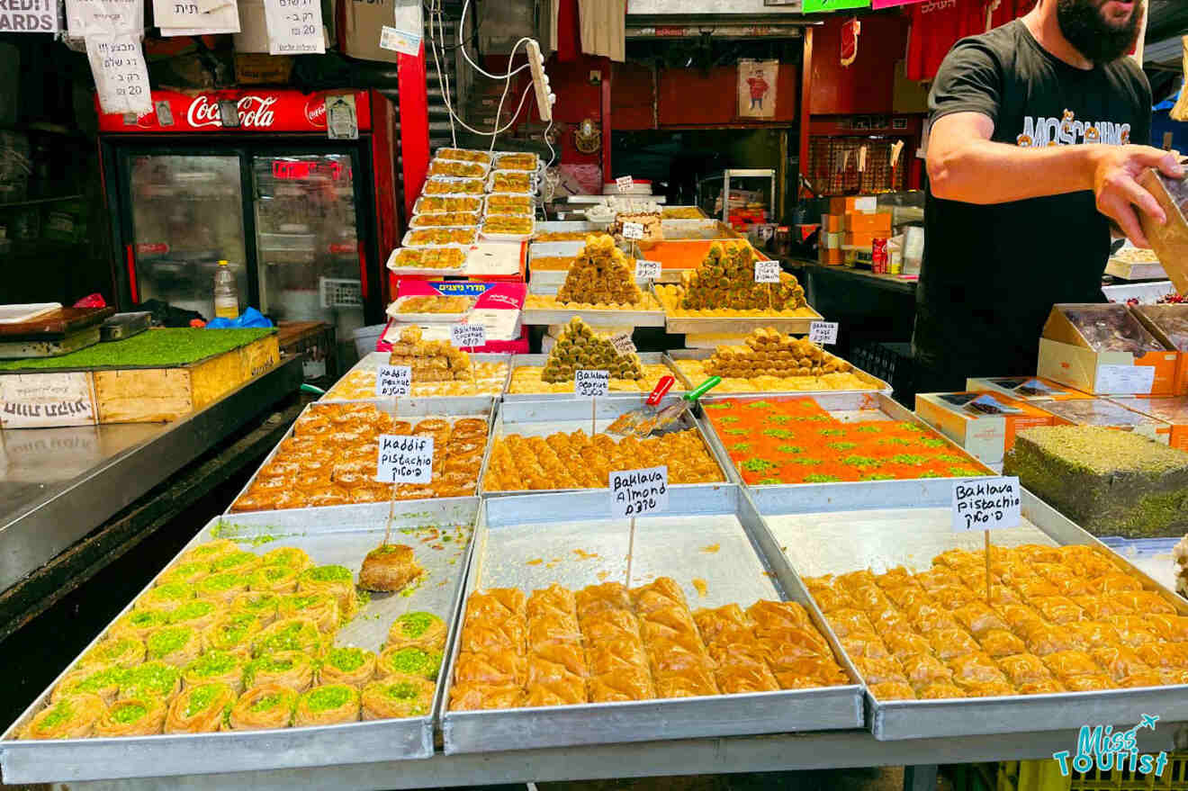 Bustling scene at Carmel Market in Tel Aviv with a variety of baklava and Middle Eastern sweets on display, and a vendor reaching out to serve the treats