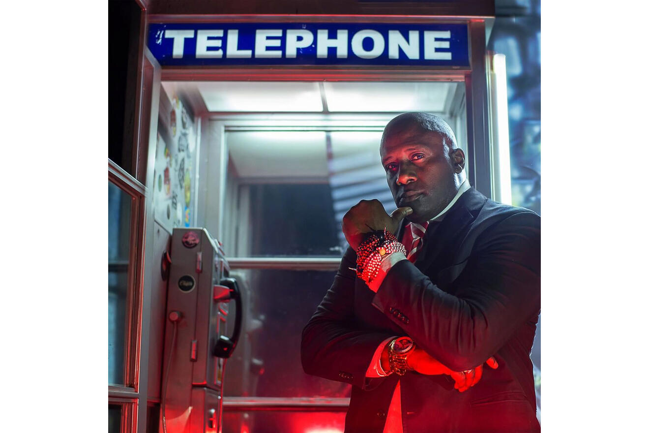 A dramatic portrait of a man in a sharp suit and red tie standing contemplatively in a red phone booth, his gaze fixed outside the frame, set against the backdrop of a dark urban night.
