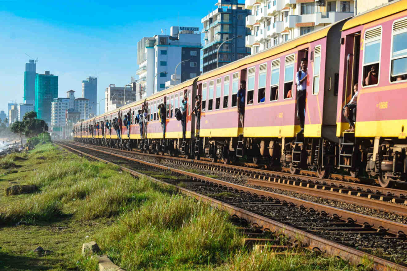 Commuters in Colombo hang onto the sides and doorways of a brightly colored train moving along the tracks, with the backdrop of a developing urban skyline under a hazy sky, capturing a dynamic moment of city transport.