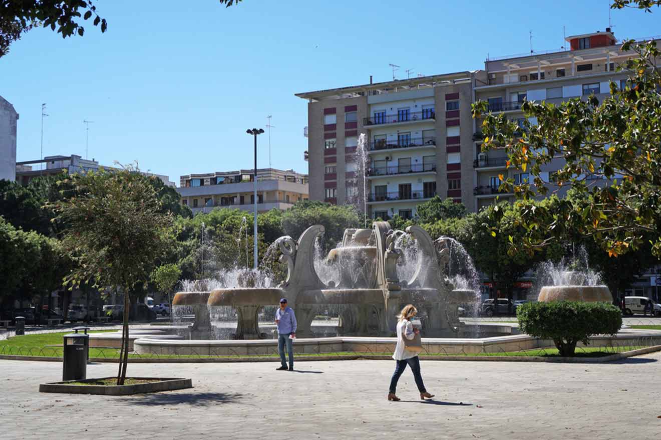"A sunny day at a spacious urban park in Lecce with people walking by a large, ornate fountain, surrounded by lush green trees and residential buildings in the background