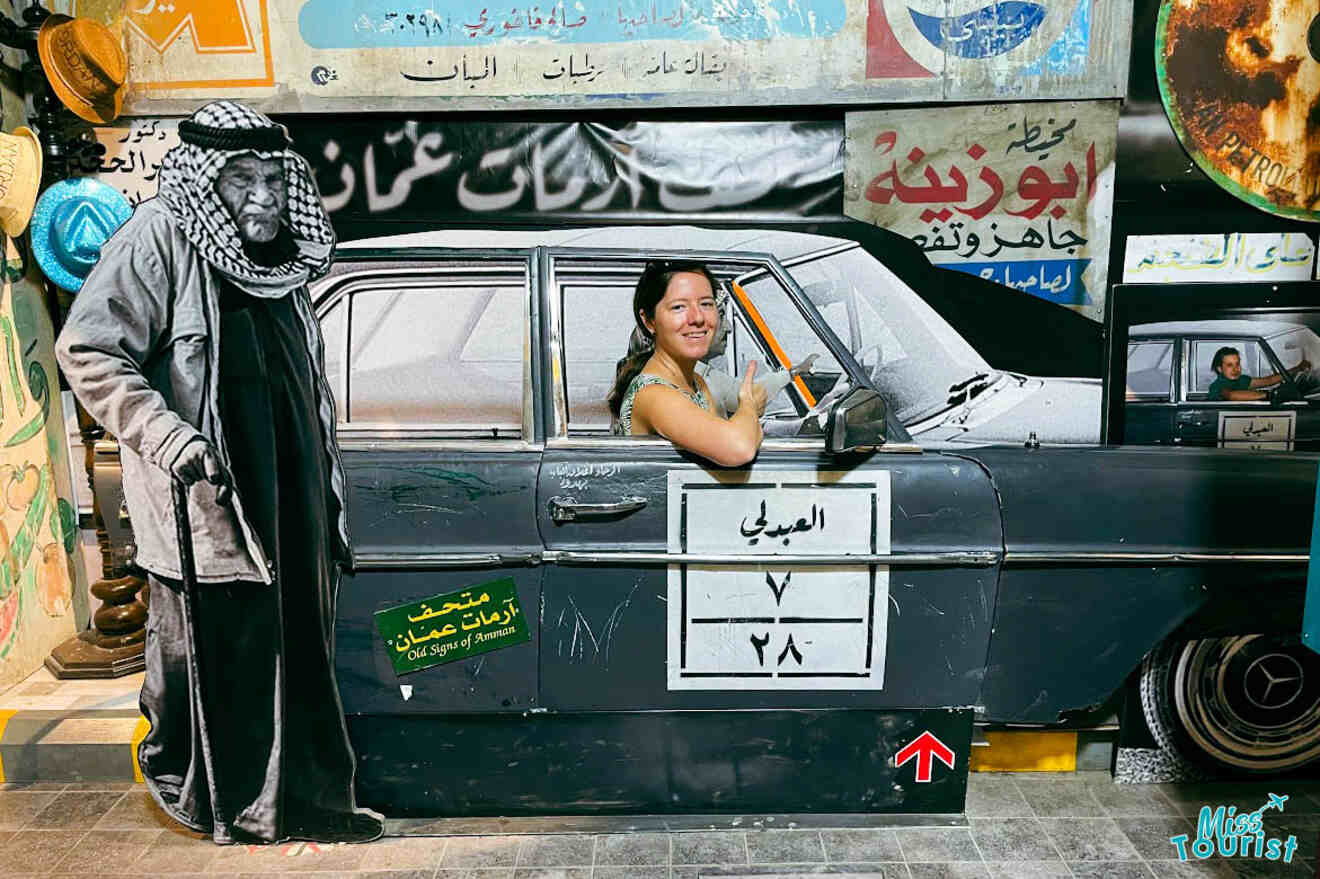 A playful photo opportunity at the Old Signs of Amman Museum with the writer of the post posing in a cutout of a vintage car alongside a life-size cardboard figure, reflecting the nostalgic charm of the museum