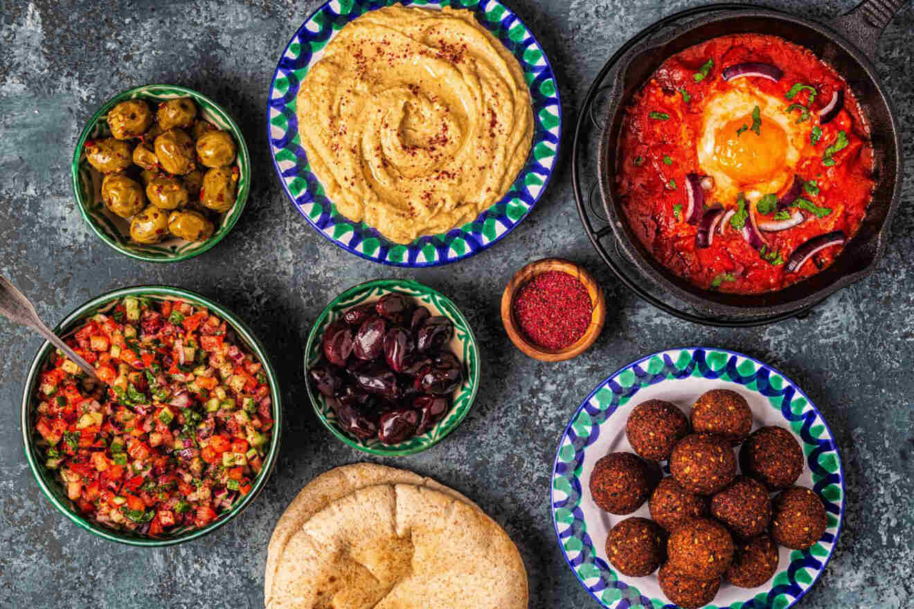 A spread of Middle Eastern cuisine including hummus, falafel, salads, olives, and pita bread, arranged on colorful plates on a textured surface