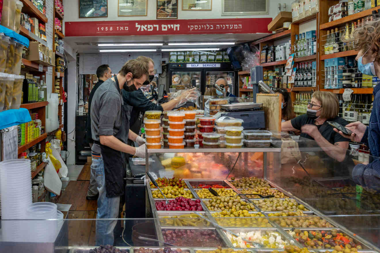 A bustling scene inside Levinsky Market in Tel Aviv with a variety of colorful pickled vegetables on display, as customers and a vendor in protective masks engage in business