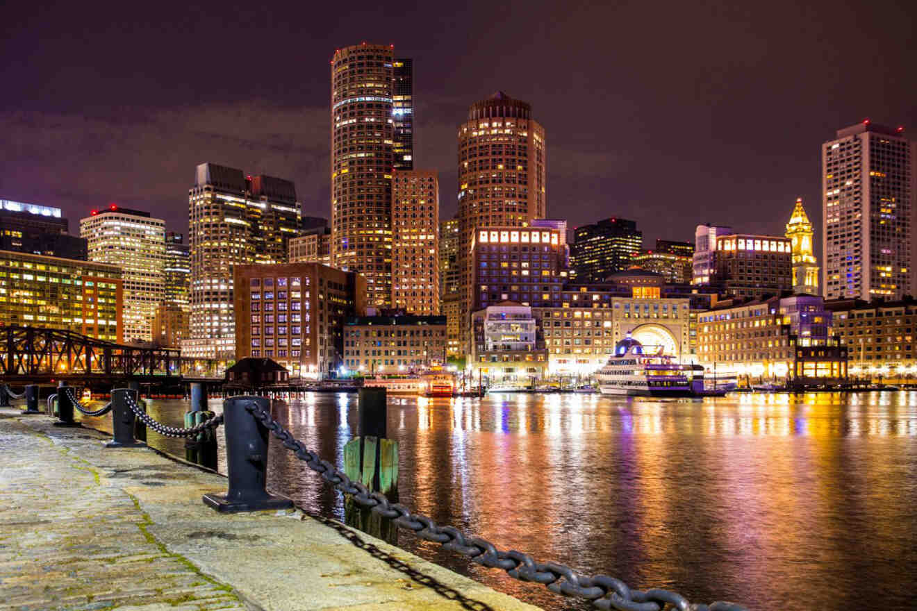 Vibrant Boston cityscape at night, showing the brightly lit buildings along the waterfront, reflecting in the water with a dock in the foreground