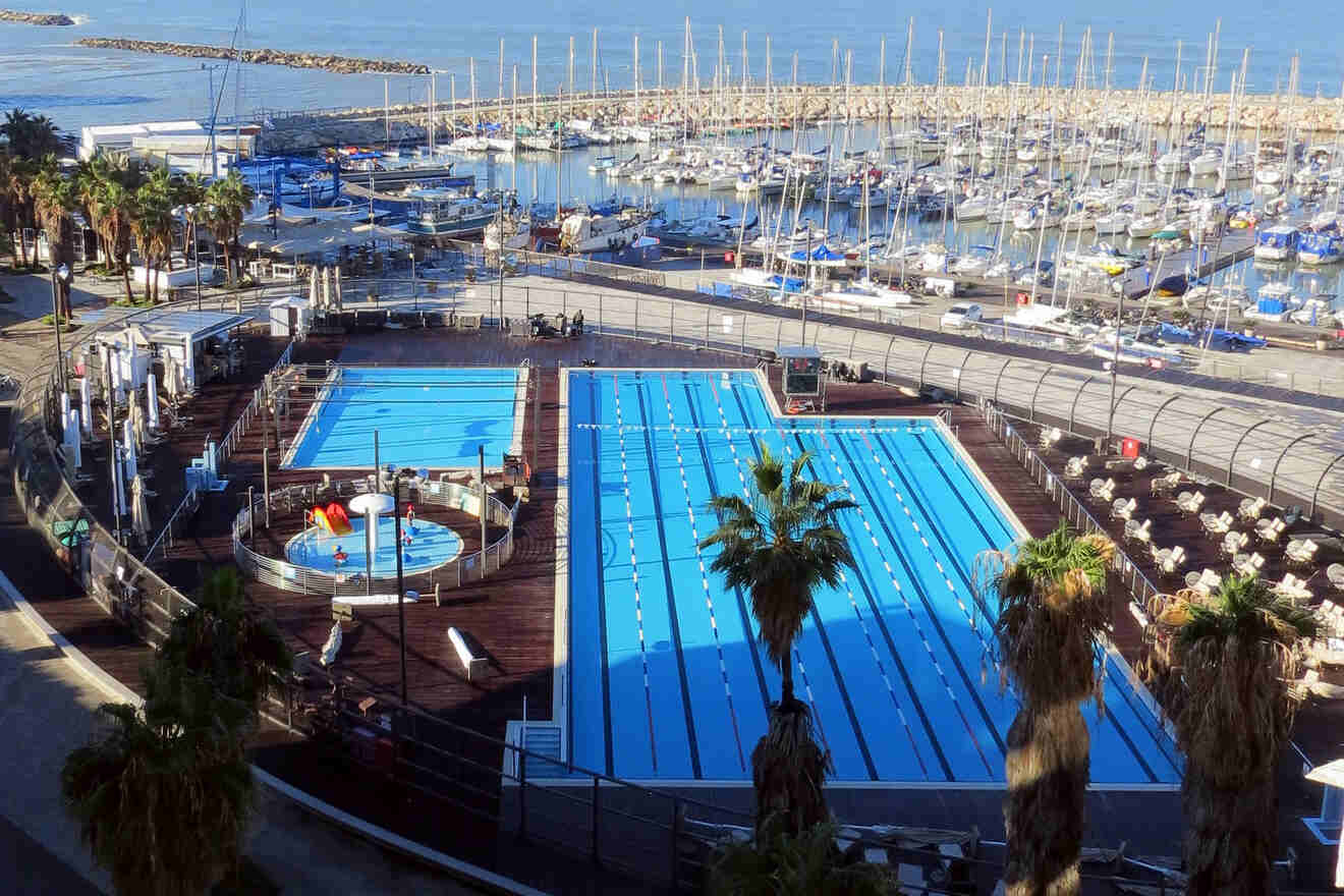 The serene Gordon Pool in Tel Aviv, with clear blue swimming lanes, surrounded by lounging areas and a vibrant marina backdrop