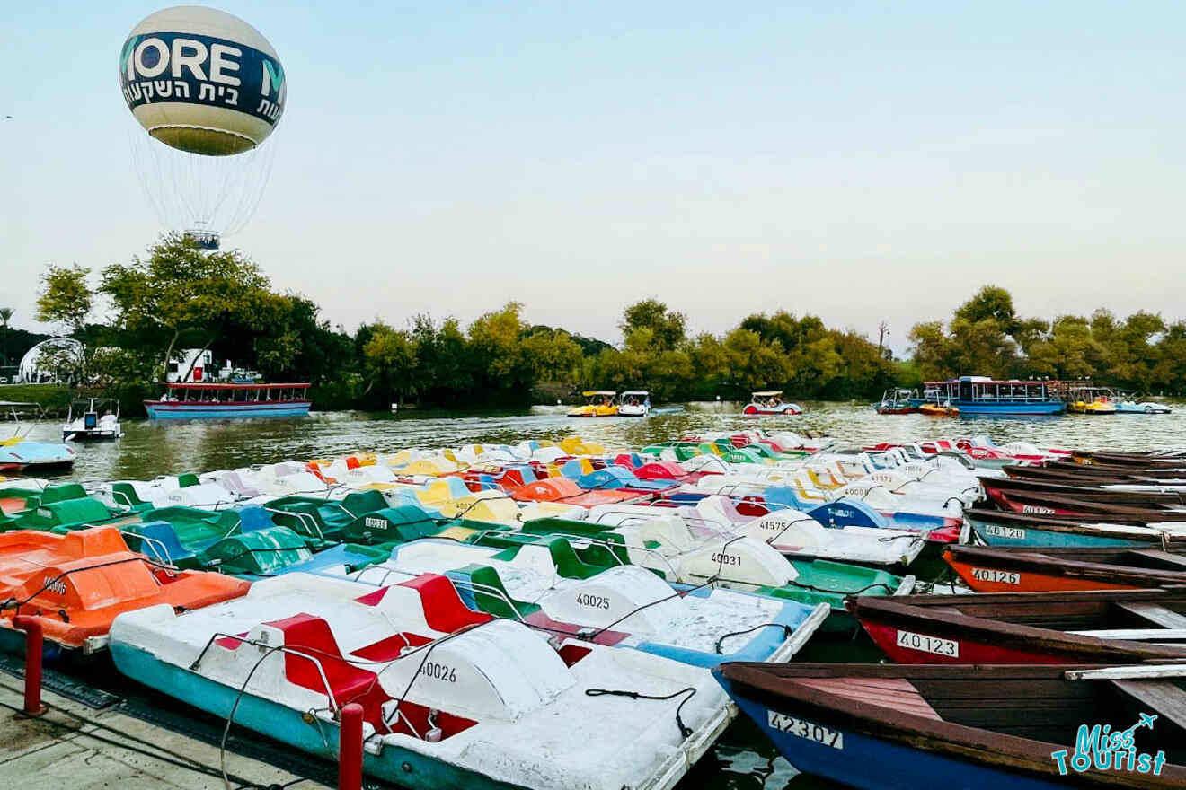 A vibrant array of pedal boats docked on the Yarkon River in HaYarkon Park, Tel Aviv, with a hot air balloon bearing 'MORE' text in the background