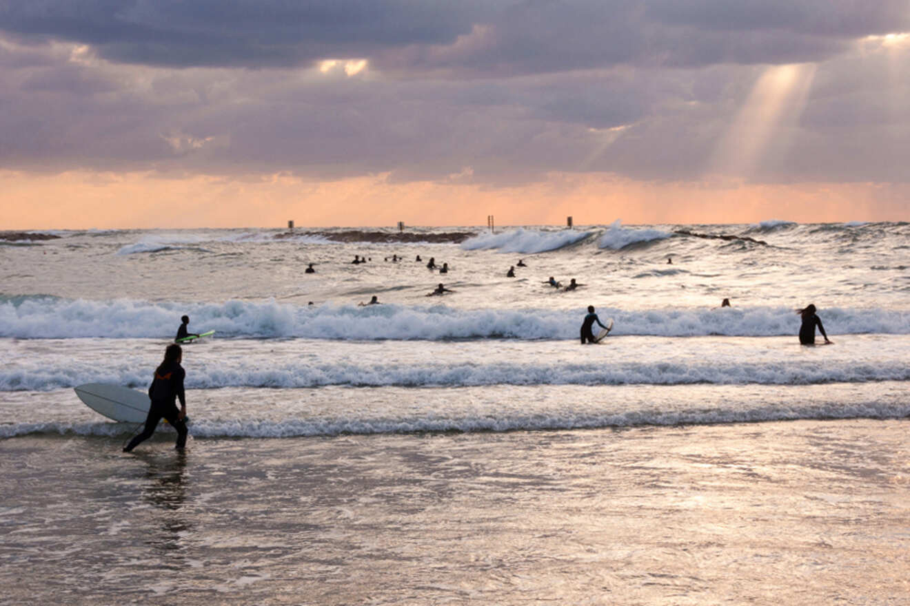 Surfers in Tel Aviv braving the wavy Mediterranean Sea at sunset, with one surfer walking out of the water carrying a surfboard under a sky with breaking sunlight through clouds