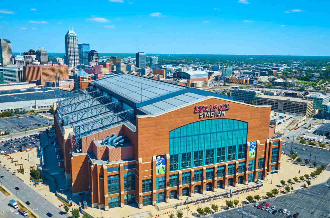 Aerial view of Lucas Oil Stadium in Indianapolis on a sunny day, featuring its large brick exterior and retractable roof with the cityscape in the background