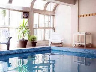An indoor pool with blue tiles, arched windows, and potted plants, creating a tranquil space for relaxation.