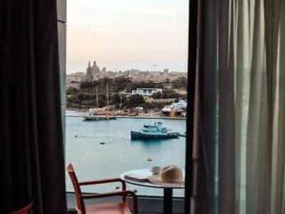 A hotel room with a balcony offering a scenic view of the harbor with boats and a cityscape in the distance, framed by curtains.