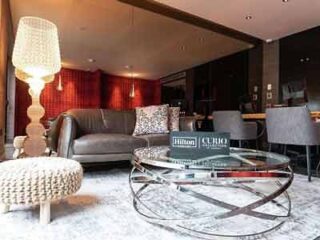 A luxurious hotel lounge with plush seating, a large floor lamp, and a glass coffee table adorned with books and a decorative piece.