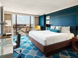 A spacious hotel room featuring a king-size bed with a vibrant blue carpet, modern furnishings, and a panoramic view of the city from the window.