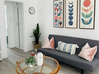 Modern living room with a gray sofa, pink and blue pillows, a round table with a plant, and abstract wall art.
