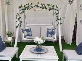 Charming outdoor swing with decorative pillows, set against a white drapery and green foliage, providing a quaint and romantic seating area.