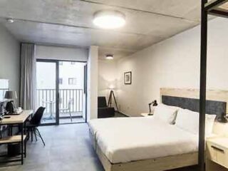 A minimalistic and modern hotel bedroom with a double bed, desk, and balcony access, featuring neutral colors.