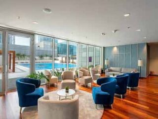 A modern hotel lounge area with sleek blue armchairs, large windows, and a view of a pool and cityscape, providing a chic relaxation space.