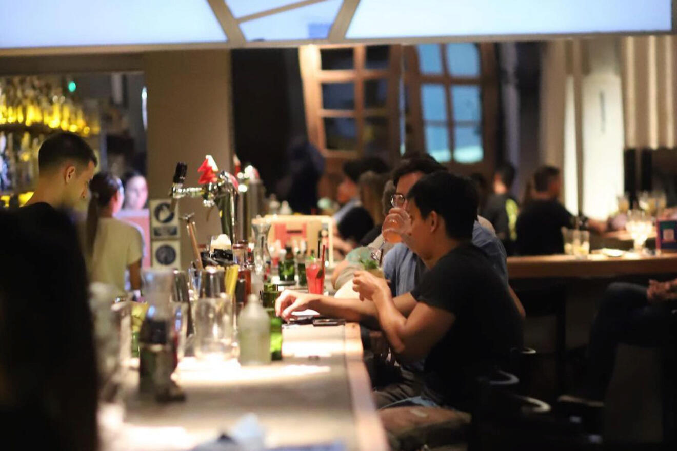 A cozy snapshot of Bar 878, where patrons engage in casual conversation while sitting at a dimly lit bar, with a bartender in the background attending to the drinks.