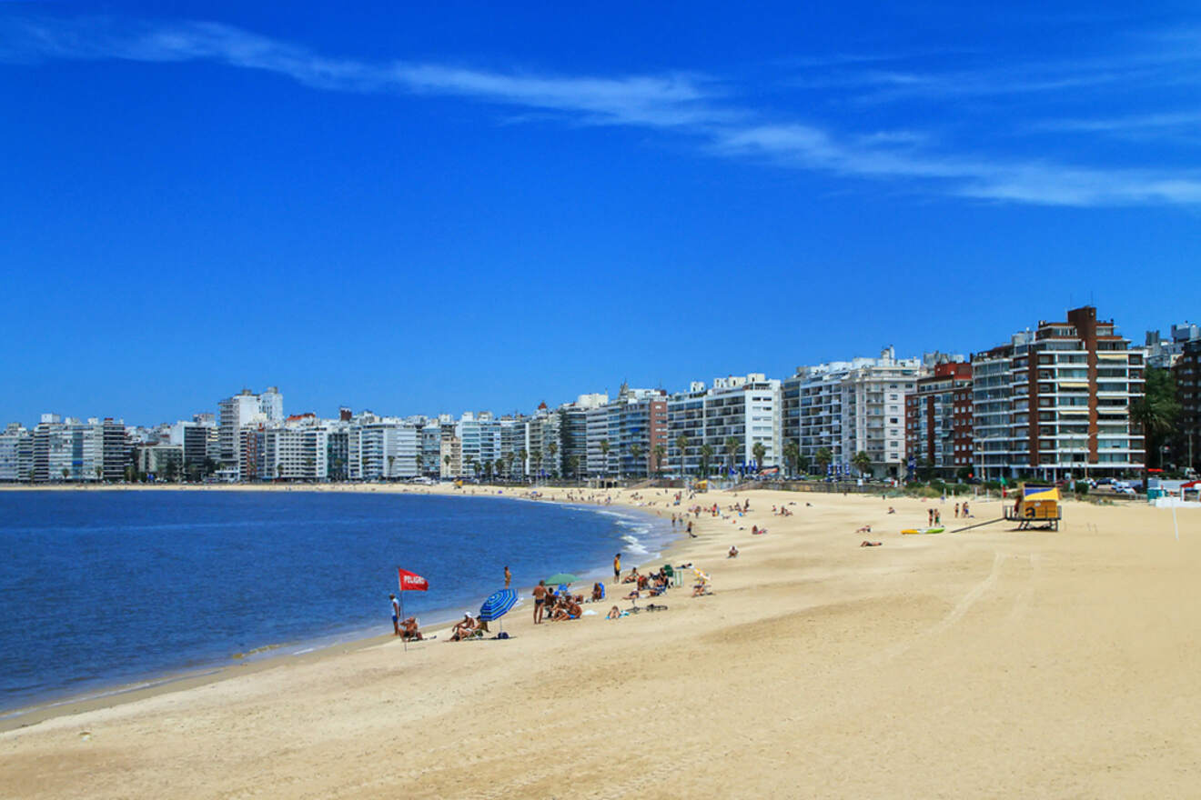 Scenic view of a sandy beach in Montevideo, Uruguay, with sunbathers and beachgoers enjoying a bright sunny day, against a backdrop of a tranquil bay and a skyline of modern buildings under a vast blue sky.