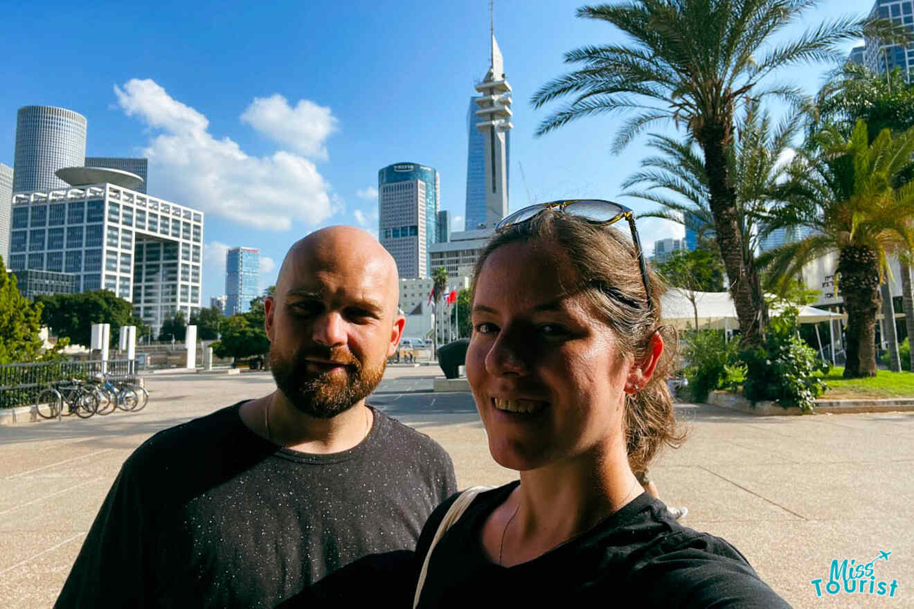 The writer and her partner taking a selfie with the modern skyline of Tel Aviv in the background, featuring skyscrapers and lush palm trees