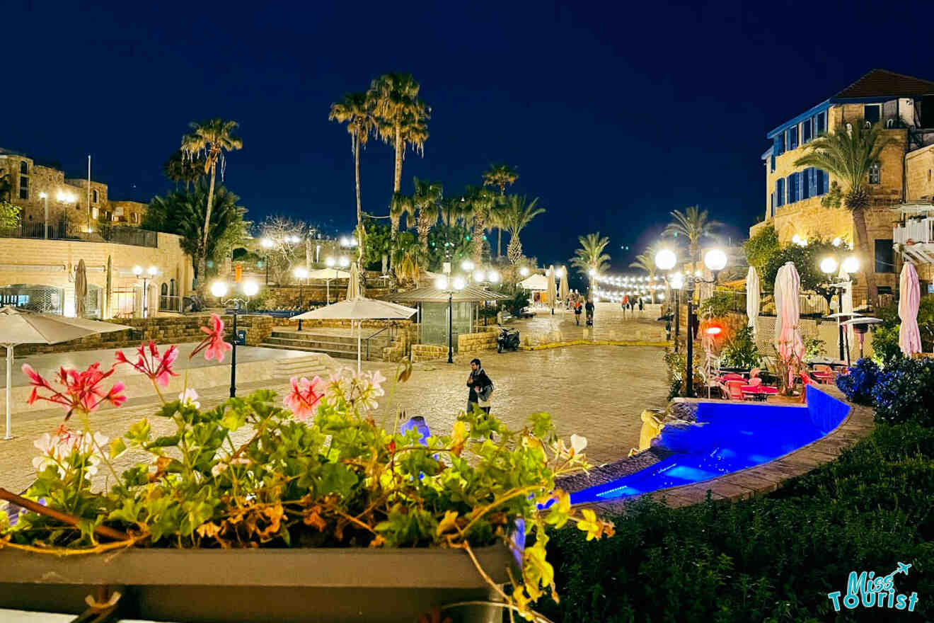 Nighttime view of a lively square in Tel Aviv, illuminated by streetlights, with palm trees, outdoor seating, and a historic building in the background